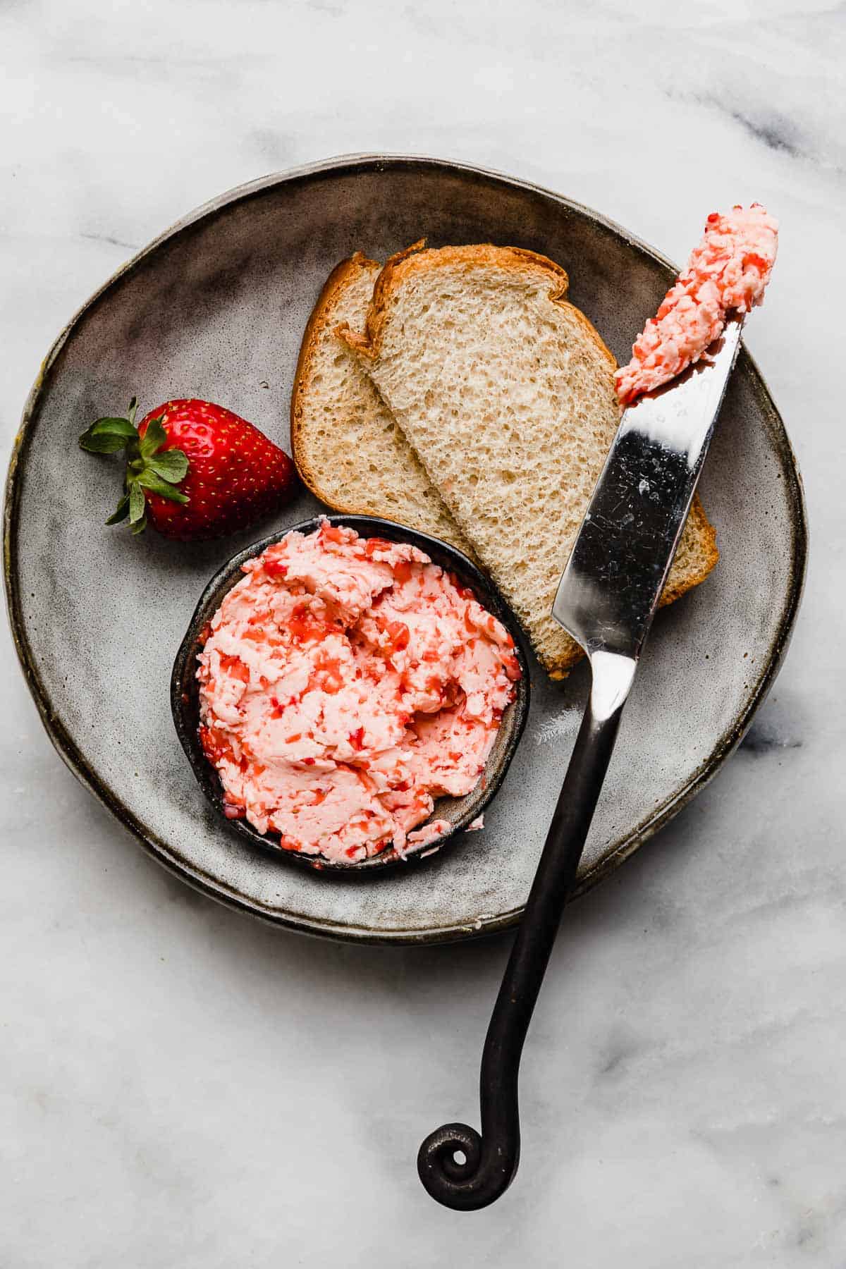 Strawberry Butter in a black bowl alongside two half slices of bread and a butter knife with fresh Strawberry Butter smeared on the knife.
