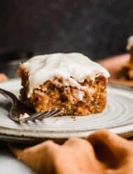 A square of Carrot Coffee Cake topped with pecan streusel and cream cheese glaze against a dark brown background.