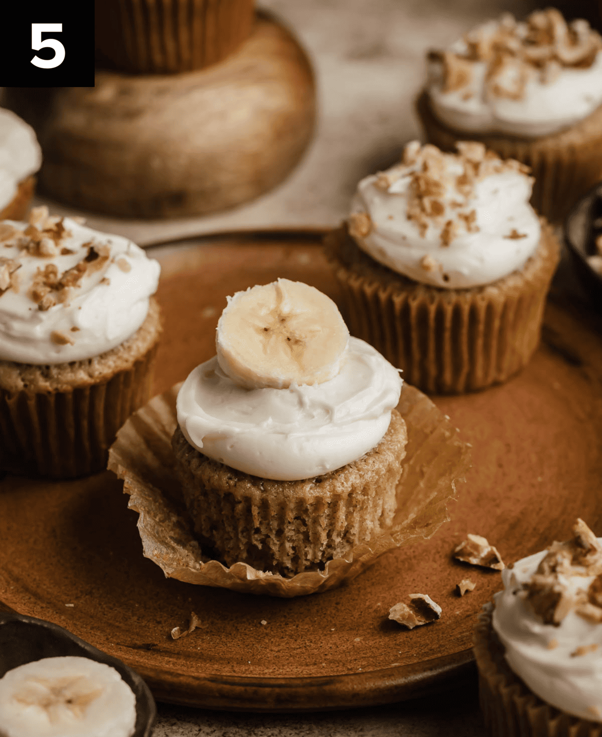 A banana cupcake topped with white cream cheese frosting with a fresh banana slice on top, the cupcake is on a brown plate.