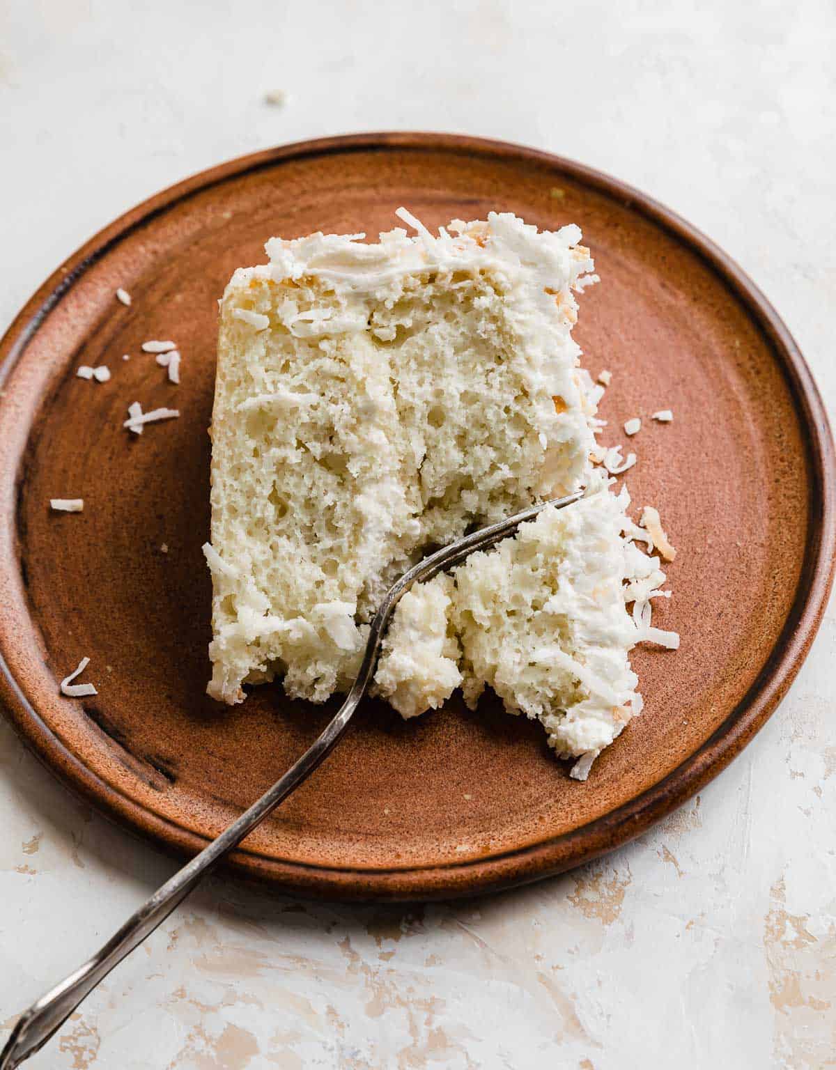 A slice of creamy Coconut Cake on a brown plate on a white background.