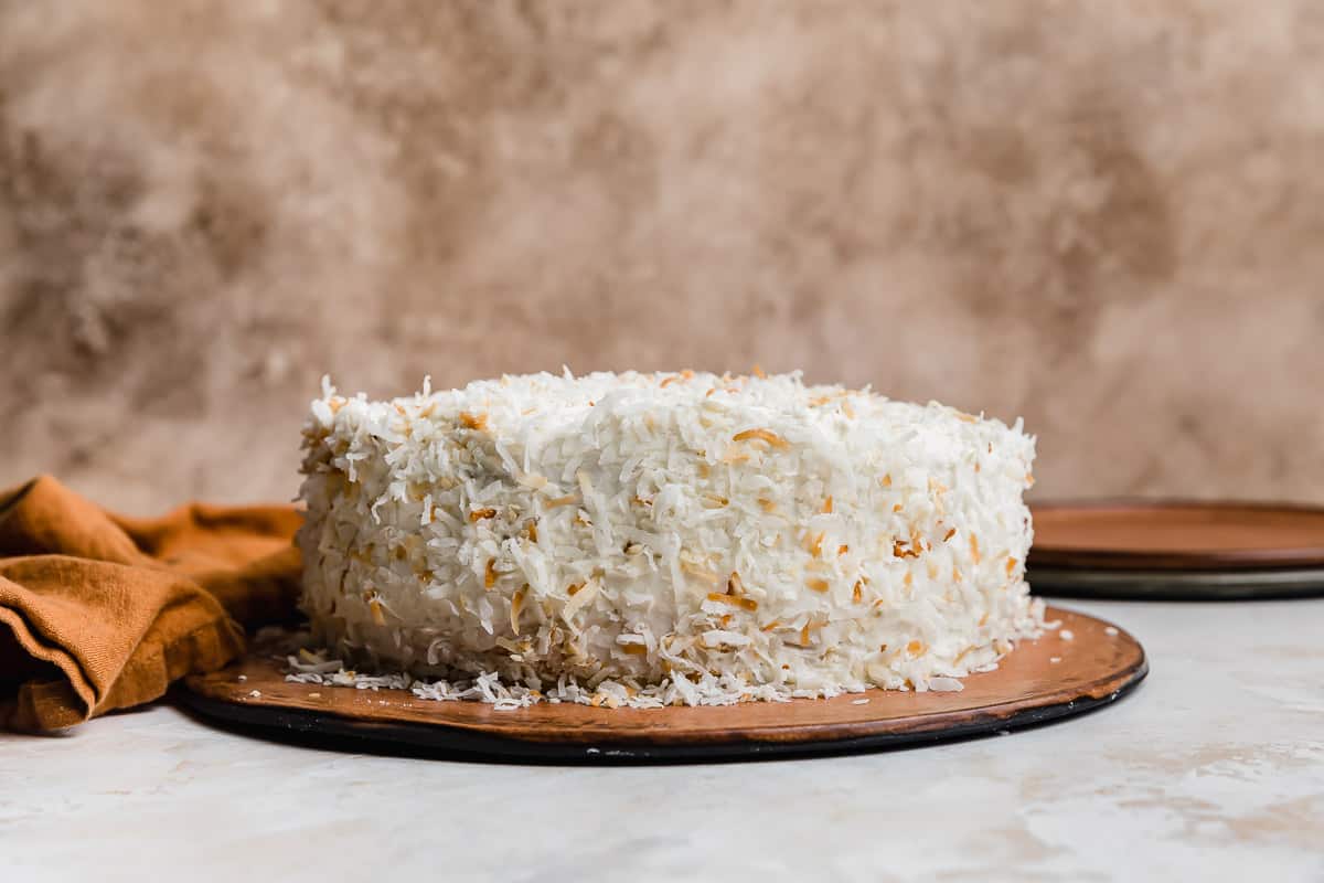 An easy coconut cake recipe on a brown cake plate topped with toasted coconut, against a light brown background.