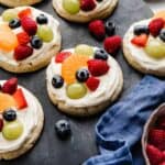 Fruit Pizza Cookies topped with a white cream cheese frosting and fresh fruit, on a black surface.