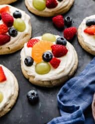 Fruit Pizza Cookies topped with a white cream cheese frosting and fresh fruit, on a black surface.