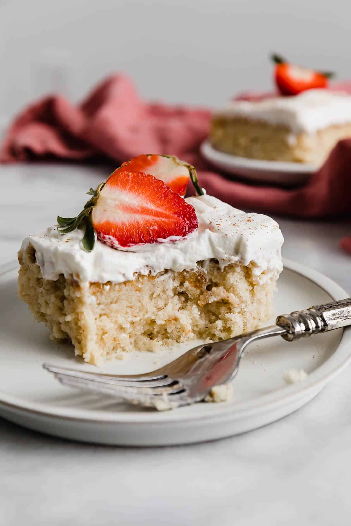 An authentic Mexican Tres Leches Cake slice on a white plate, topped with a slice of fresh strawberries, with a bite taken out of the tres leches cake. 