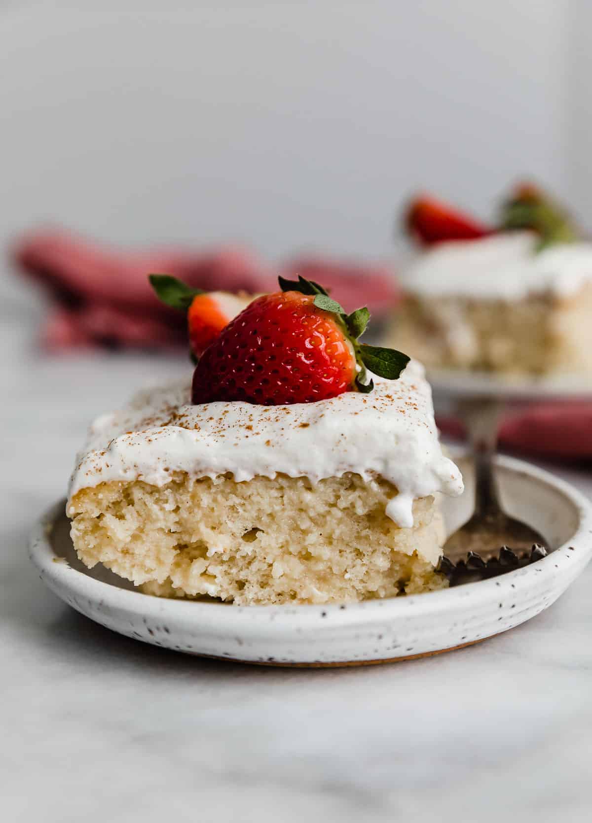 A slice of Tres Leches Cake on a white plate, with a fresh strawberry on the whipped cream.