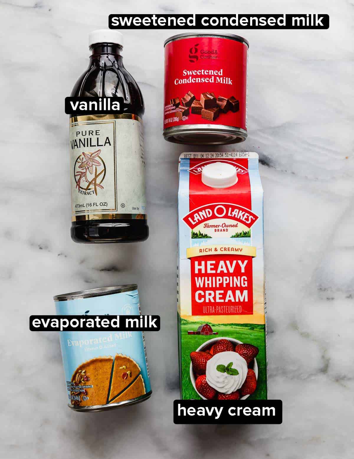 Three milks (evaporated milk, sweetened condensed milk, and heavy cream) paired with a bottle of vanilla extract on a marble surface.