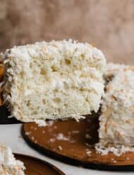 A black spatula balancing a slice of coconut cake with coconut frosting and toasted coconut.