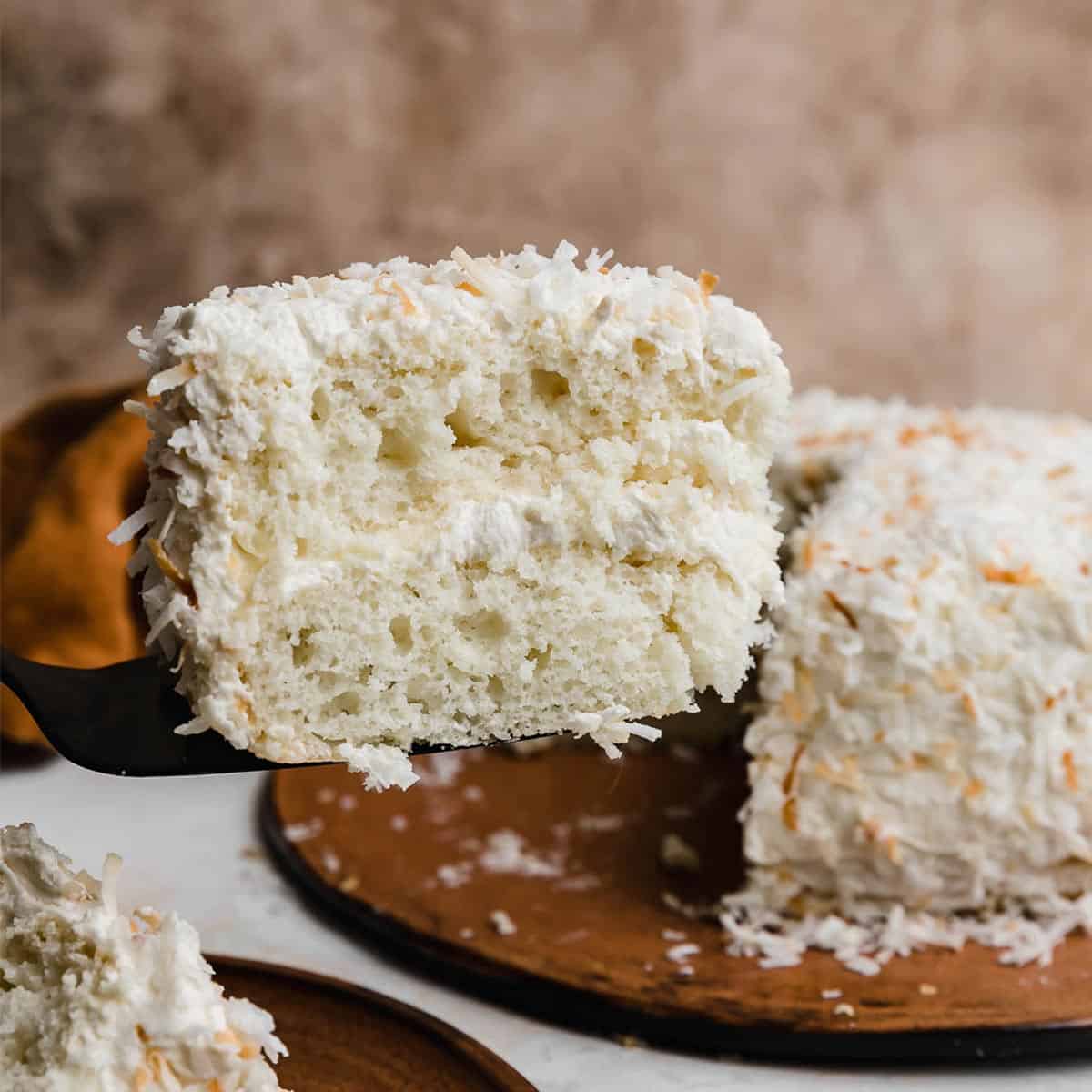 A black spatula balancing a slice of coconut cake with coconut frosting and toasted coconut.
