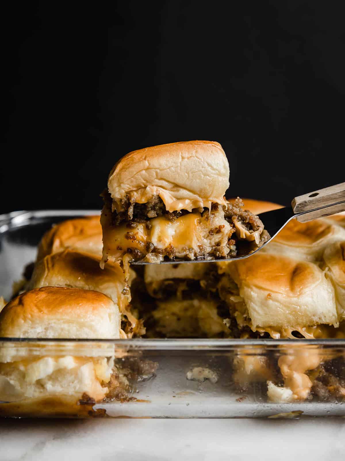 A Hawaiian Roll Breakfast Sliders recipe being held up on a metal spatula against a black background.