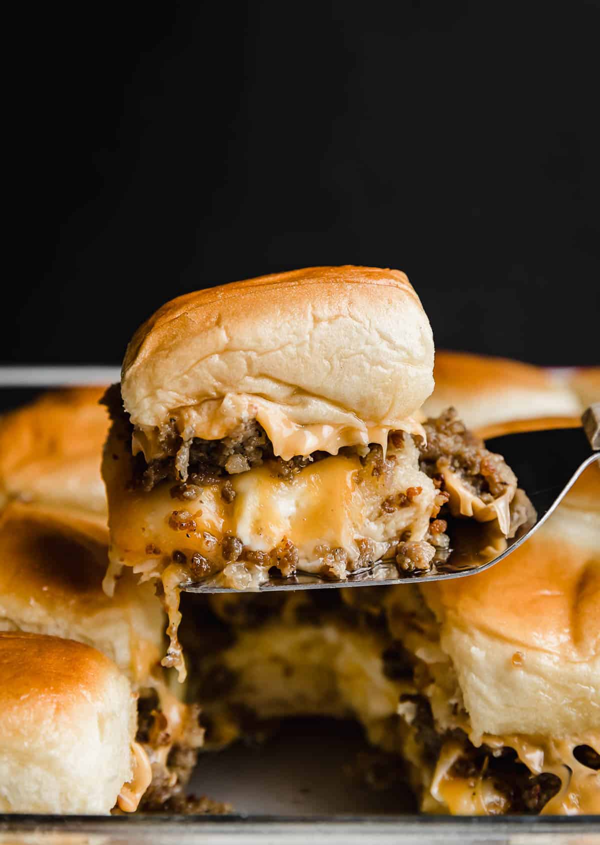 Easy Breakfast Sliders with sausage and cheesy scrambled eggs made on Hawaiian rolls, balancing on a metal spatula against a black background.