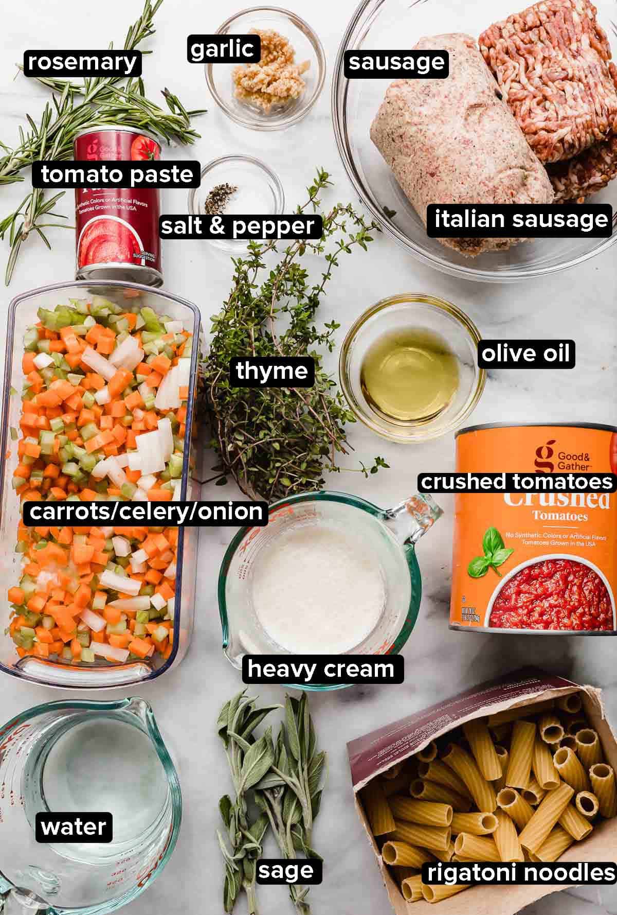 Sausage Ragu ingredients on a white marble surface: fresh herbs such as sage, thyme, rosemary, crushed tomatoes, carrots, onion, celery, sausage and Italian sausage, rigatoni noodles, heavy cream, and tomato paste.