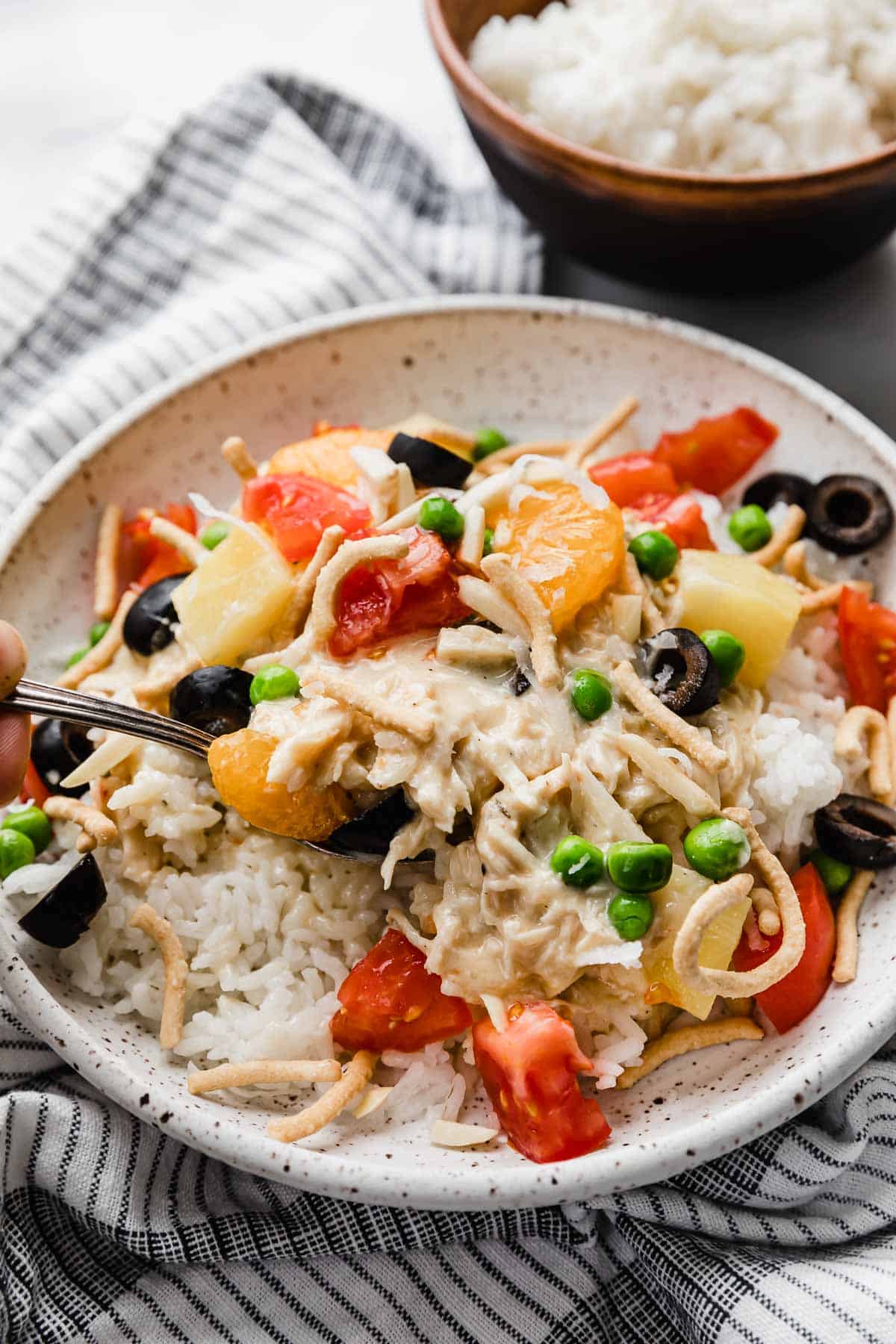 A white plate loaded with the best Hawaiian Haystacks recipe and toppings including tomatoes, pineapple, coconut, olives, and chow mein noodles.