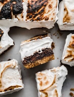 S'mores Brownies cut into squares, with one S'more brownie on its side showcasing the graham cracker crust, fudgy brownie, and homemade toasted marshmallow cream topping.