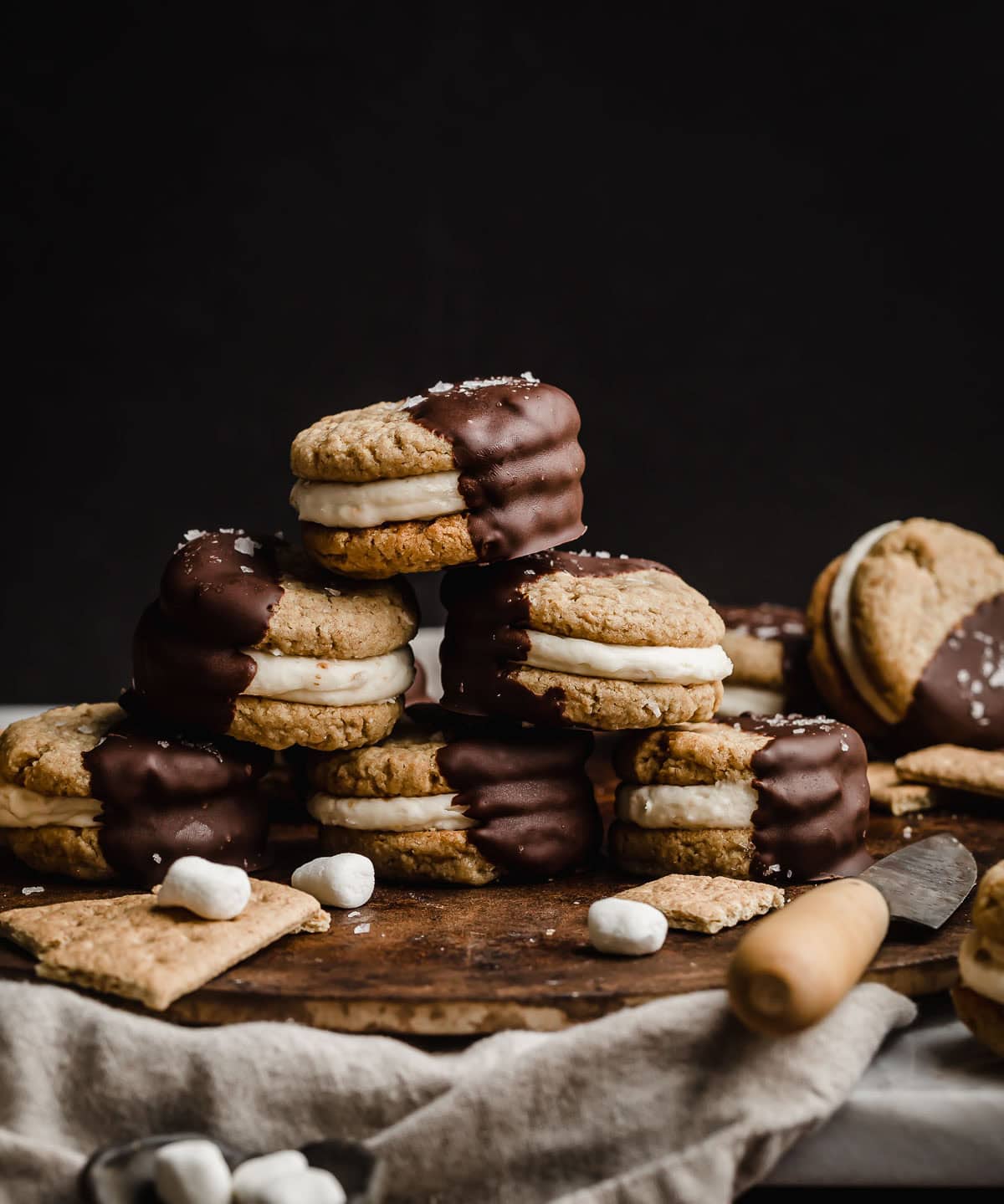 S'mores Sandwich Cookies with marshmallow frosting between graham cracker cookies then half dipped in chocolate, against a black background.