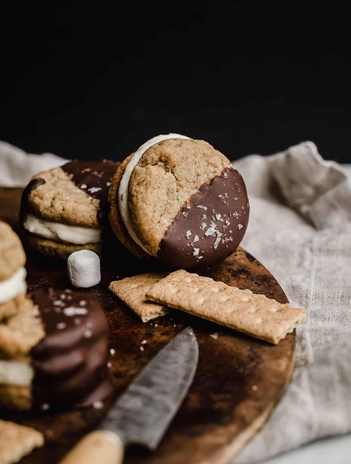 A s'mores cookie sandwich with marshmallow frosting and dipped in melted chocolate, leaning against a brown surface.