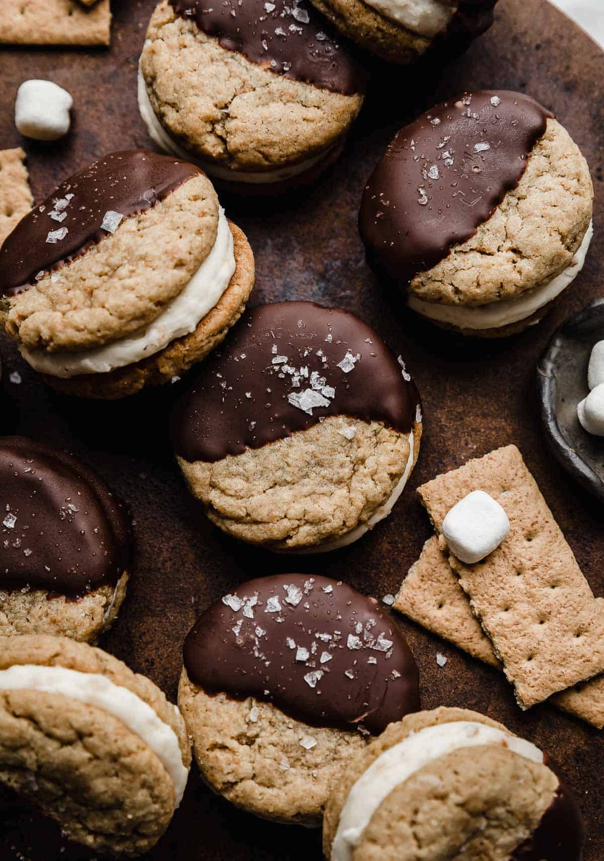 Several of the best S'mores Sandwich Cookies on a dark brown surface.