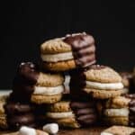 S'mores Sandwich Cookies halfway dipped in melted chocolate against a dark black background.