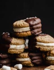 S'mores Sandwich Cookies halfway dipped in melted chocolate against a dark black background.