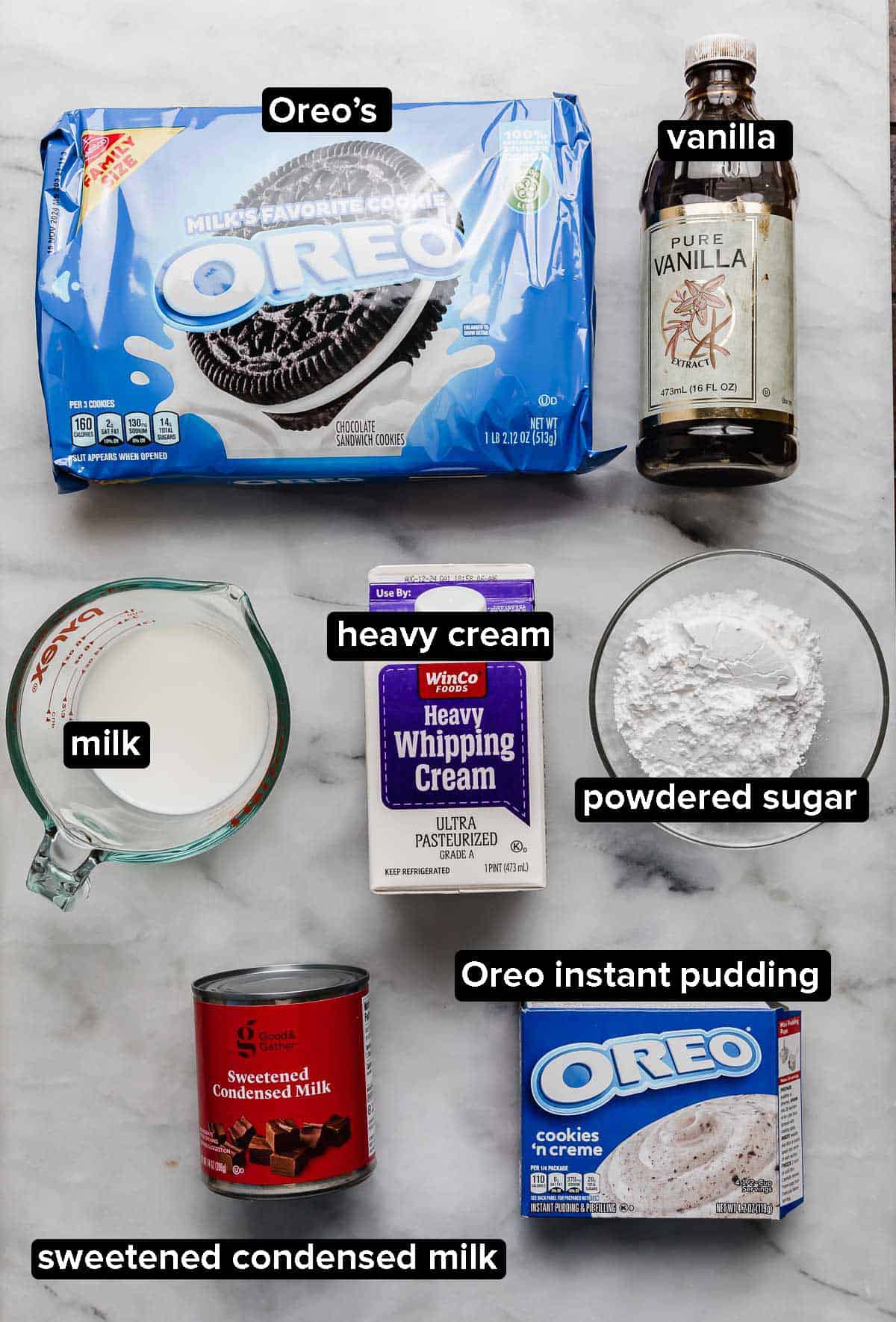 Oreo Poke Cake frosting ingredients on a white surface: package of Oreo cookies, vanilla, heavy cream, milk, Oreo instant pudding, sweetened condensed milk, and powdered sugar.