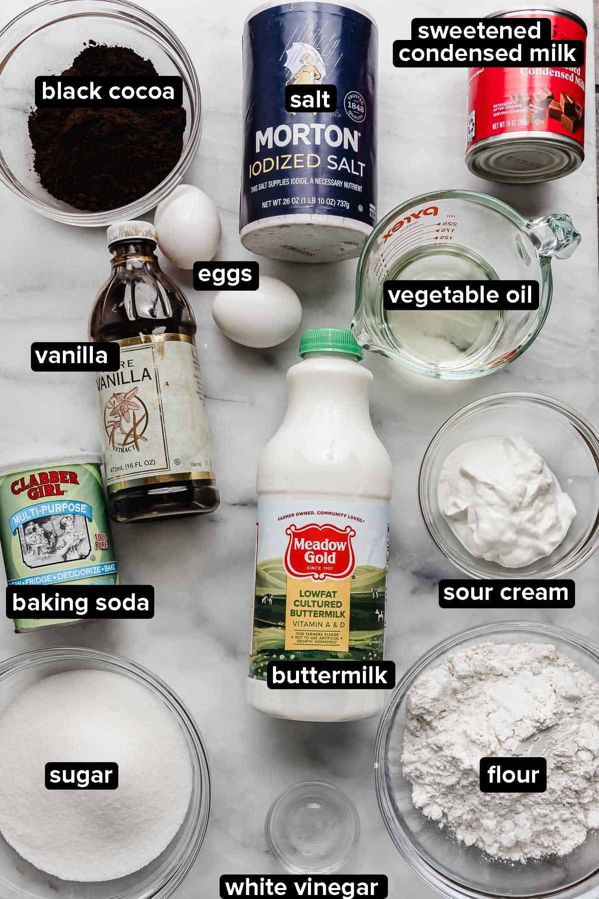 Oreo Poke Cake recipe ingredients on a white marble surface, ingredients include black cocoa, buttermilk, sour cream, flour, salt, eggs, oil, and flour.