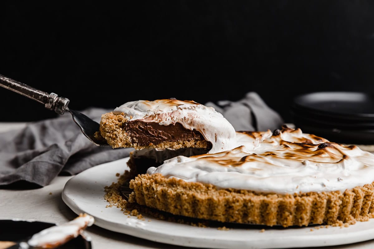 A slice of homemade S'mores Pie balanced on a pie spatula hovering against a black background.