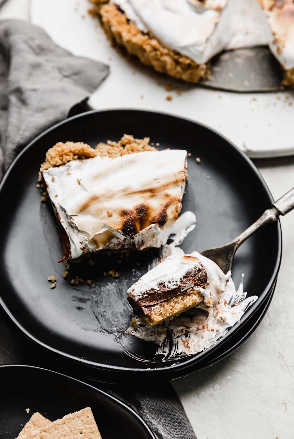 A slice of S'mores Pie on a black plate with a fork cutting into a chunk.