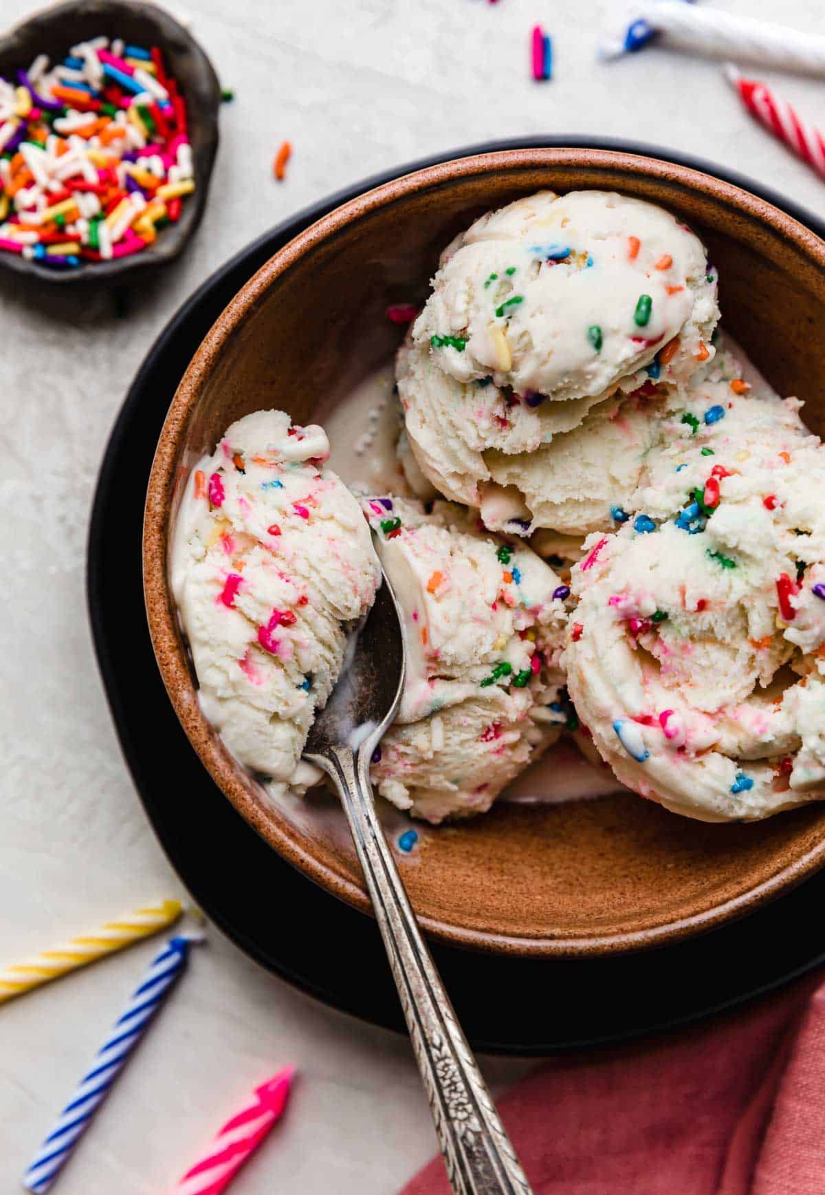 Three scoops of Birthday Cake Ice Cream with sprinkles, in a brown bowl.