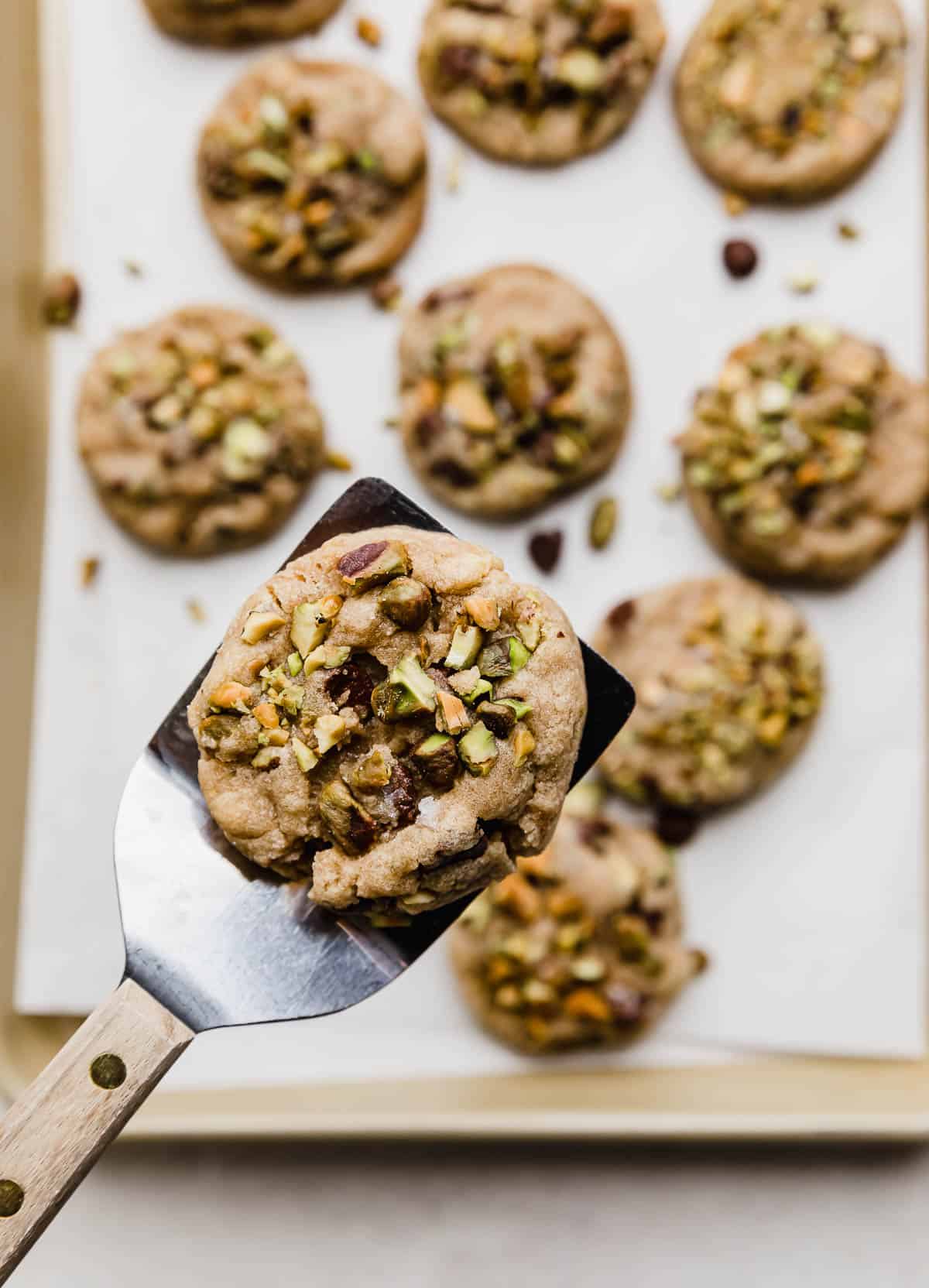 A Chocolate Chip Pistachio Cookie balancing on a metal spatula hovering over a baking sheet of the best pistachio chocolate chip cookies.