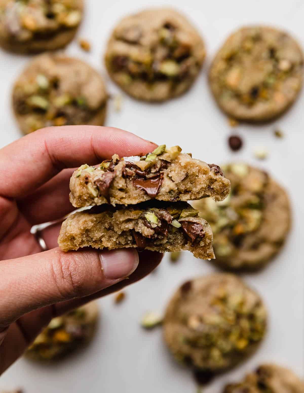 A hand holding a chocolate chip pistachio cookie that's been broken in half.