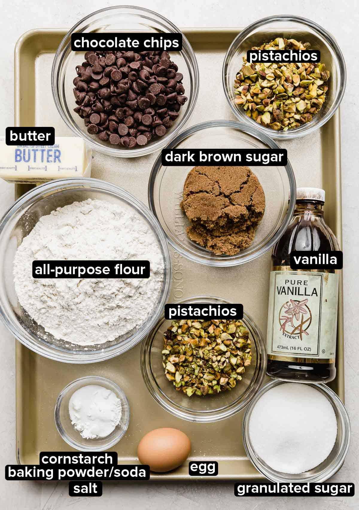 Chocolate Chip and Pistachio Cookie ingredients portioned into glass bowls on a baking sheet: egg, flour, chocolate chips, dark brown sugar, vanilla, pistachios, and butter.