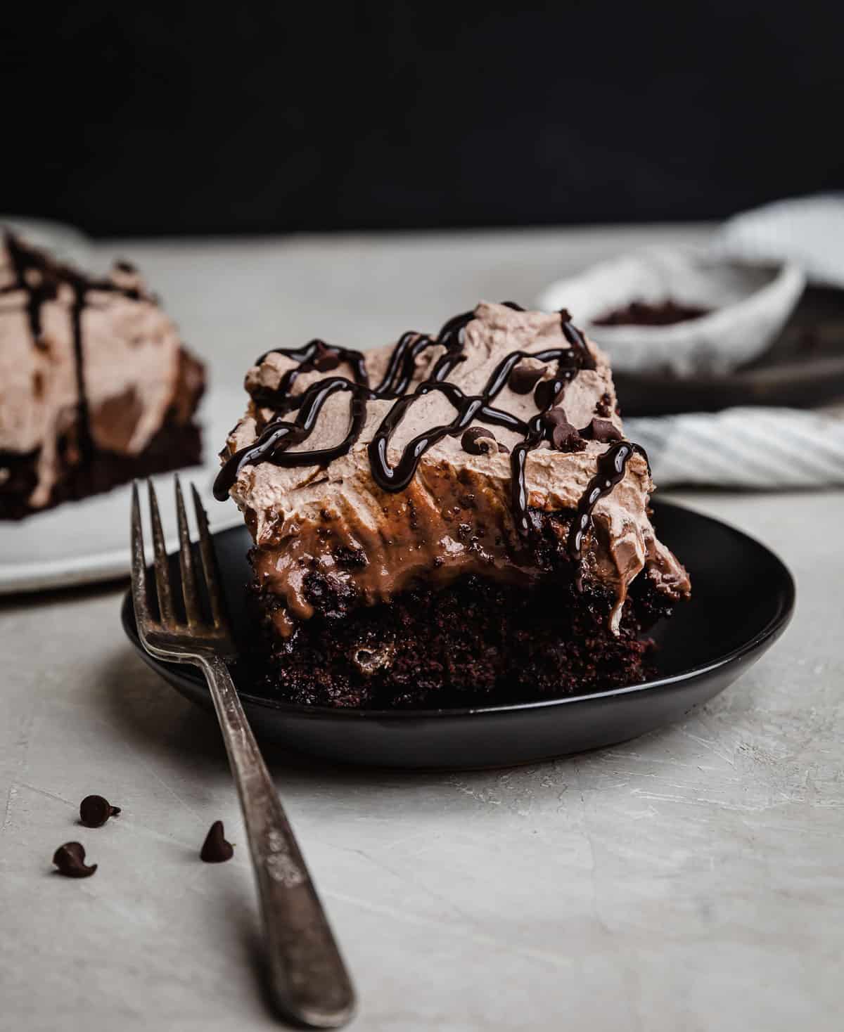 Chocolate pudding cake (Chocolate Poke Cake recipe) on a black plate, topped with whipped cream and chocolate sauce.