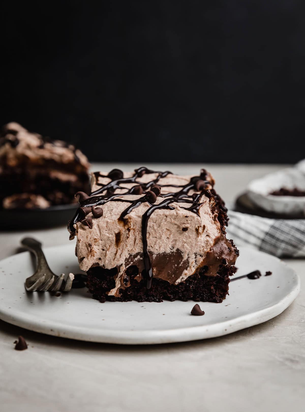 A Chocolate Poke Cake with chocolate pudding and chocolate whipped cream topping on a white plate against a black background.