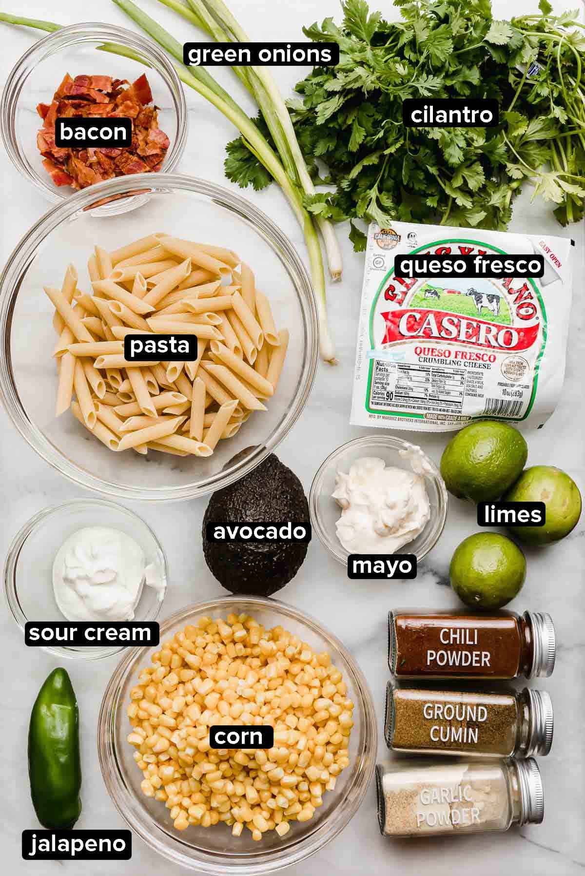 Mexican Street Corn Pasta Salad ingredients on a white marble surface: pasta, cilantro, jalapeño, corn, bacon, avocado, queso fresco, limes, and seasonings. 