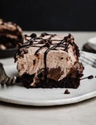 Chocolate Poke Cake topped with chocolate pudding, chocolate whipped cream, and drizzle of chocolate sauce, on a white plate that's against a black background.