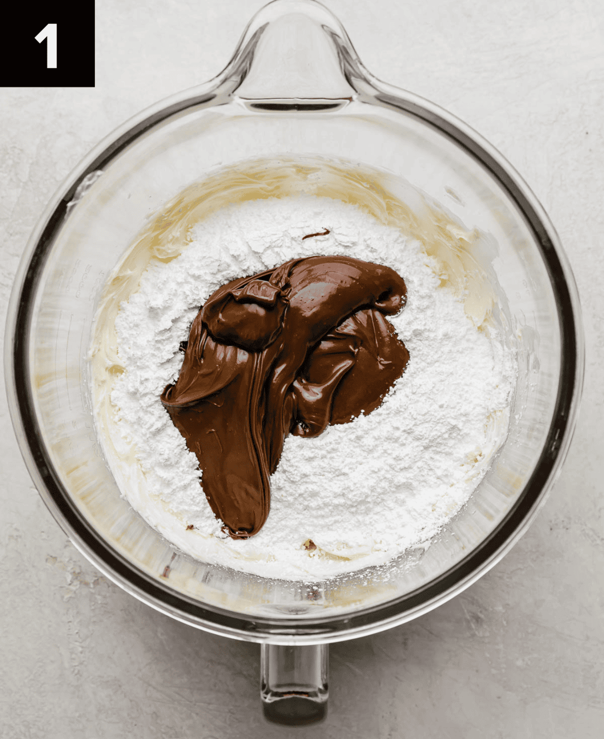 A glass mixing bowl with butter, powdered sugar, and Nutella in it (on a white background).