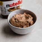 Hazelnut spread frosting (Nutella frosting recipe) in a white bowl with a jar of Nutella in the background.