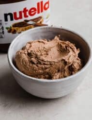 Hazelnut spread frosting (Nutella frosting recipe) in a white bowl with a jar of Nutella in the background.