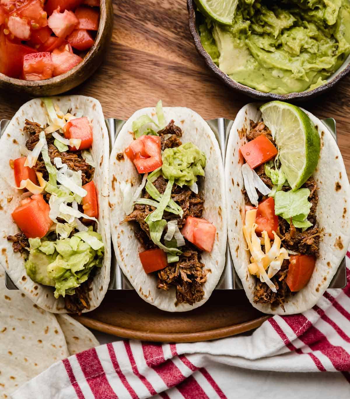 Three flour tortillas filled with Crock Pot Shredded Beef Taco mix, shredded lettuce, guacamole, and tomatoes.