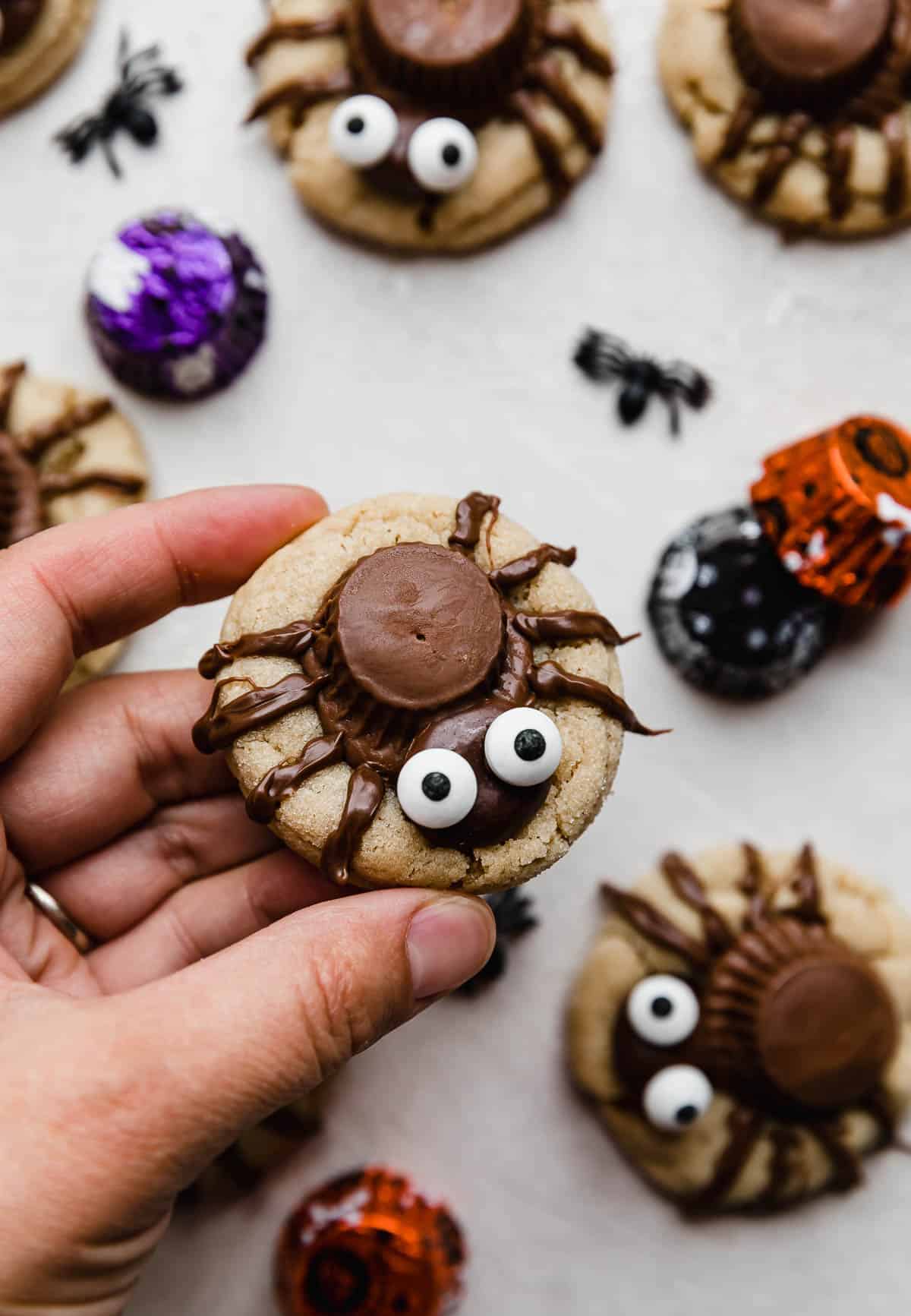 A hand holding a Spider cookie made with a peanut butter cookie base, upside down Reese's peanut butter cup, milk dud topped with candy eyes, and melted chocolate piped spider legs.