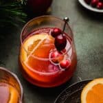 A cranberry orange mocktail in a glass cup on a green surface with a slice of orange and fresh cranberries garnishing the drink.