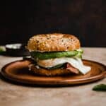 An Everything Bagel Breakfast Sandwich with egg, bacon, spinach, avocado and cream cheese on a brown plate against a black background.
