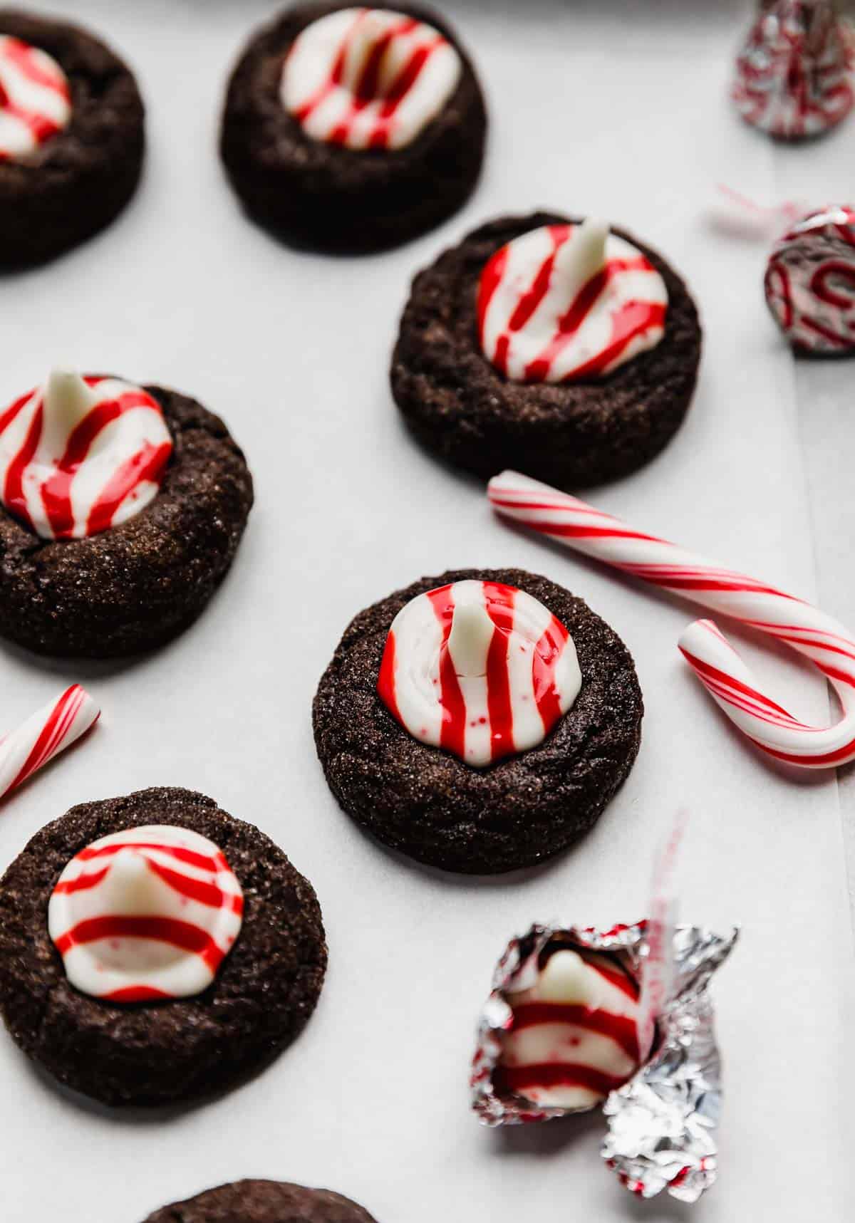 A chocolate cookie topped with a red and white striped peppermint candy kiss, on a white surface with a small candy cane next to the Christmas blossom cookie.