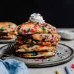 A stack of Birthday Cake Pancakes with whipped cream on top against a black background.