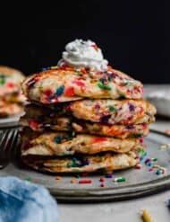 A stack of Birthday Cake Pancakes with whipped cream on top against a black background.