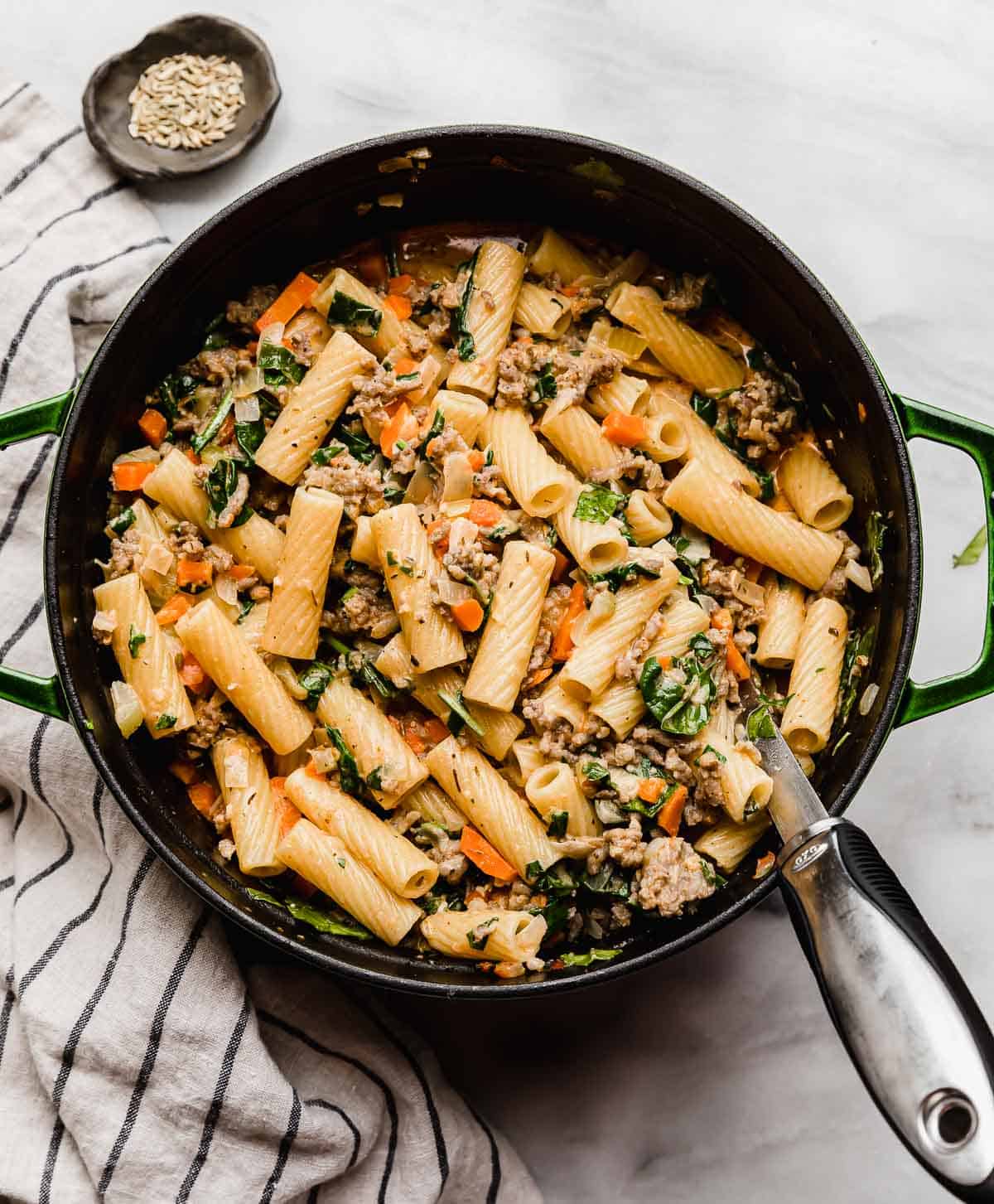 A green dutch oven filled with Italian Sausage Rigatoni with spinach on a white marble surface.