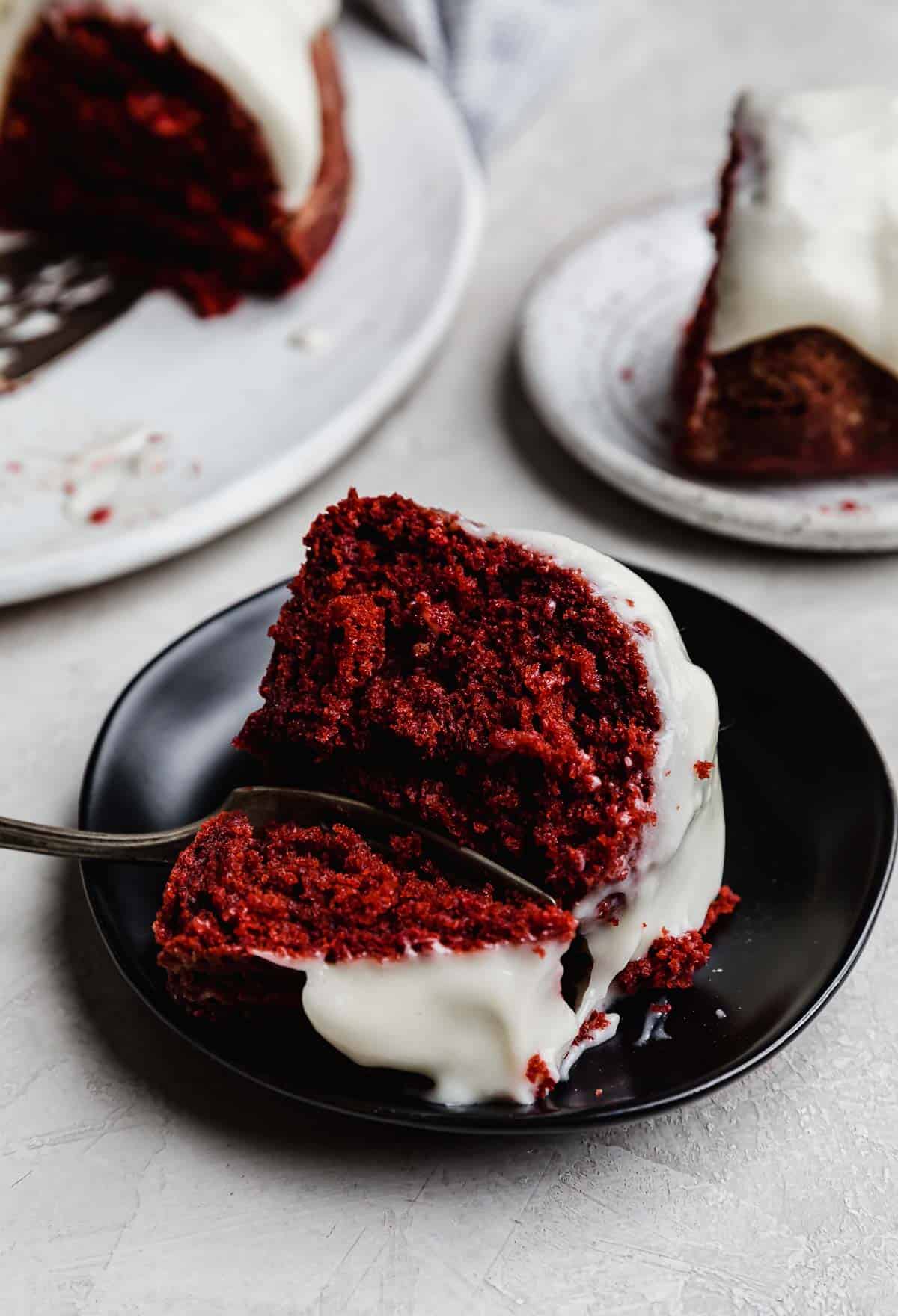 A slice of Red Velvet Bundt Cake topped with glaze on a black plate.