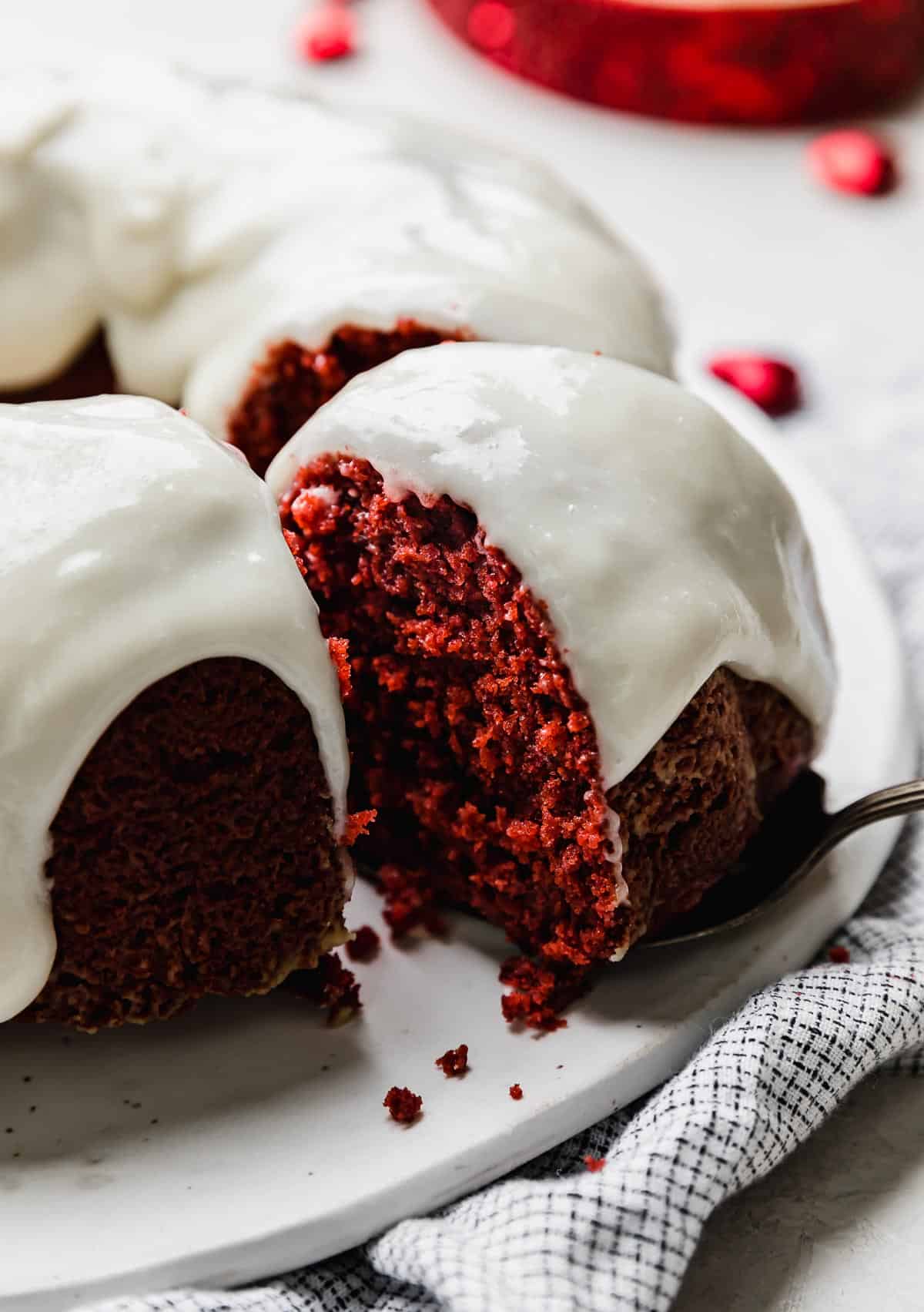 A moist Red Velvet Bundt Cake topped with cream cheese glaze and a slice of cake partially removed from the red bundt cake.