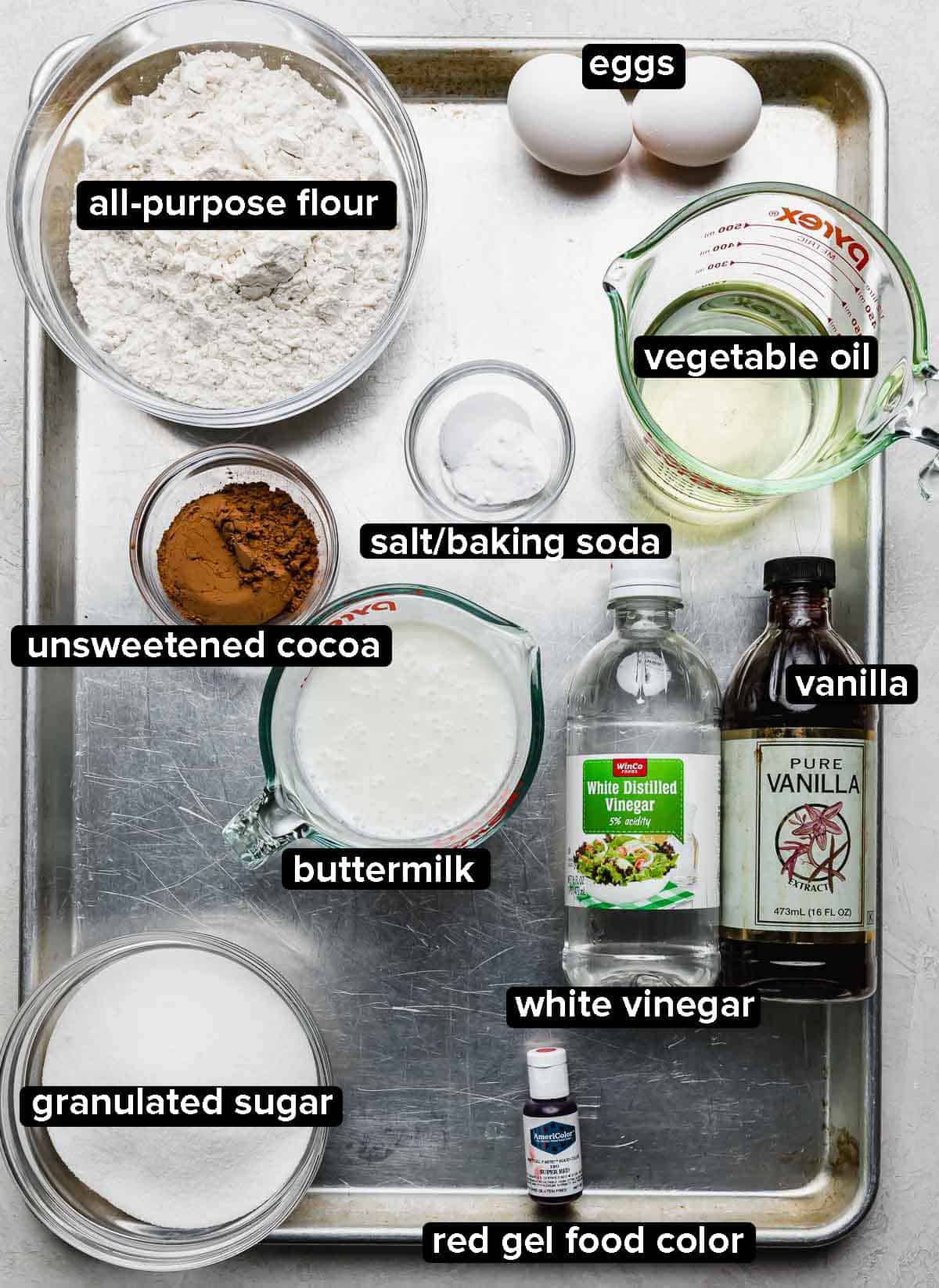 Red Velvet Bundt Cake ingredients portioned into glass bowl on a silver baking sheet: oil, buttermilk, vanilla, red gel food color, sugar, cocoa powder, eggs, salt and baking soda, and white vinegar.