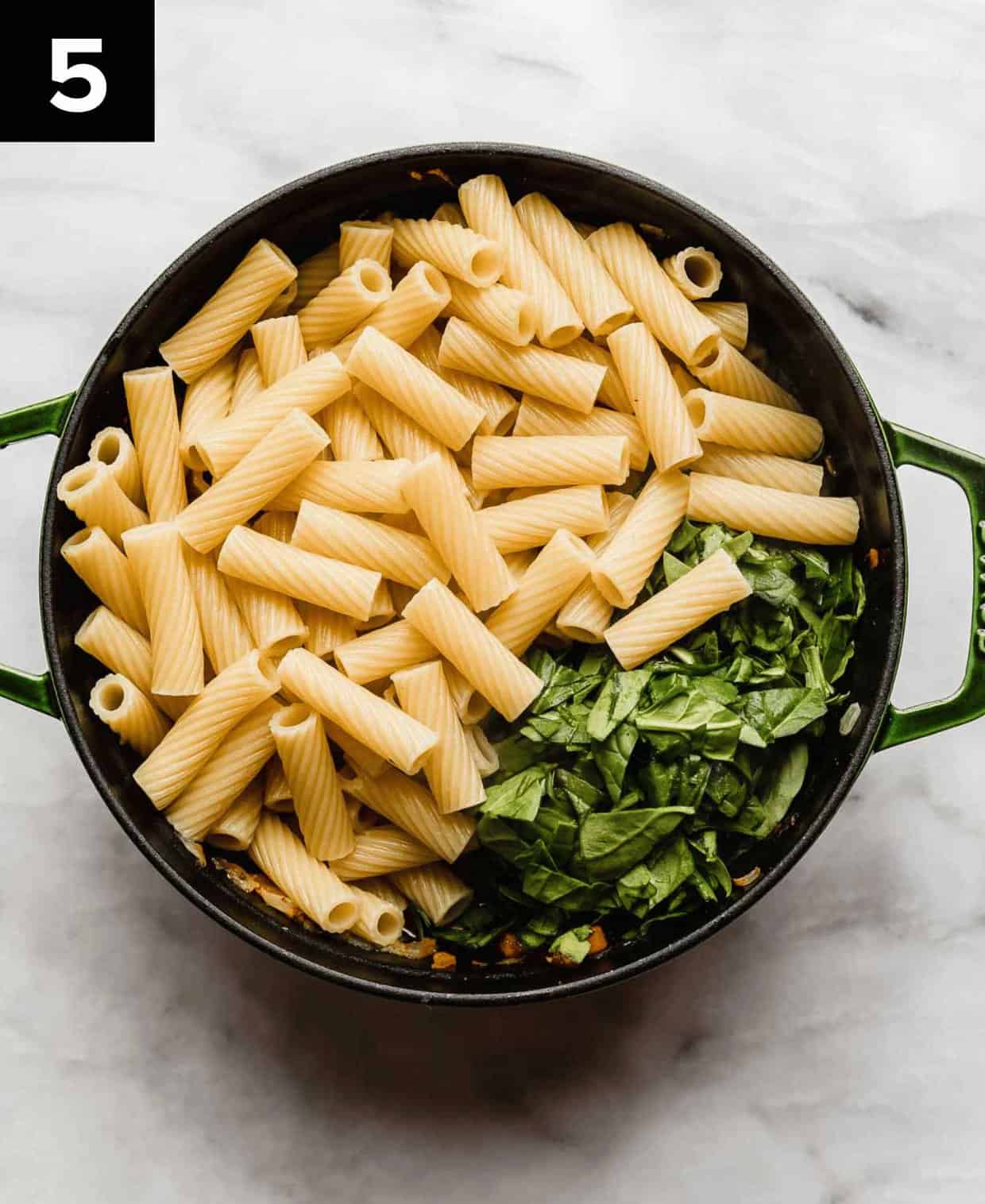 Cooked rigatoni noodles and chopped spinach in a large dutch oven on a white surface.