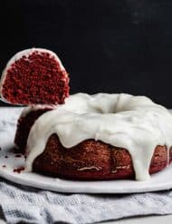 A slice of red valentines day bundt cake hovering over a cream cheese glaze topped Red Velvet Bundt Cake against a black background.